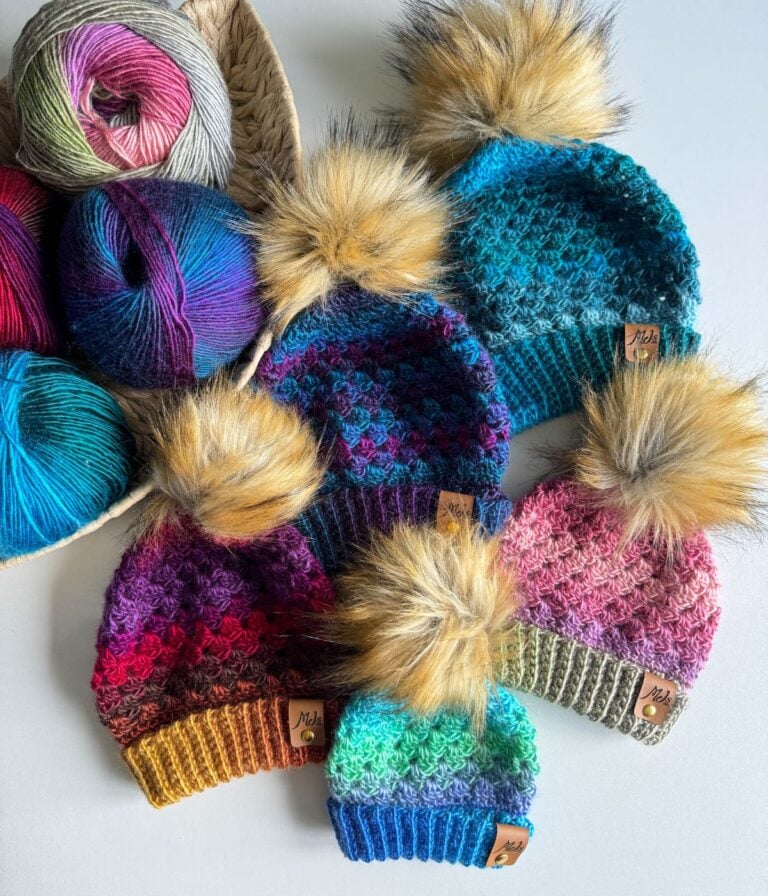 A collection of colorful Granny Stitch hats with faux fur pom-poms, arranged on a table, next to several skeins of yarn in a woven basket.