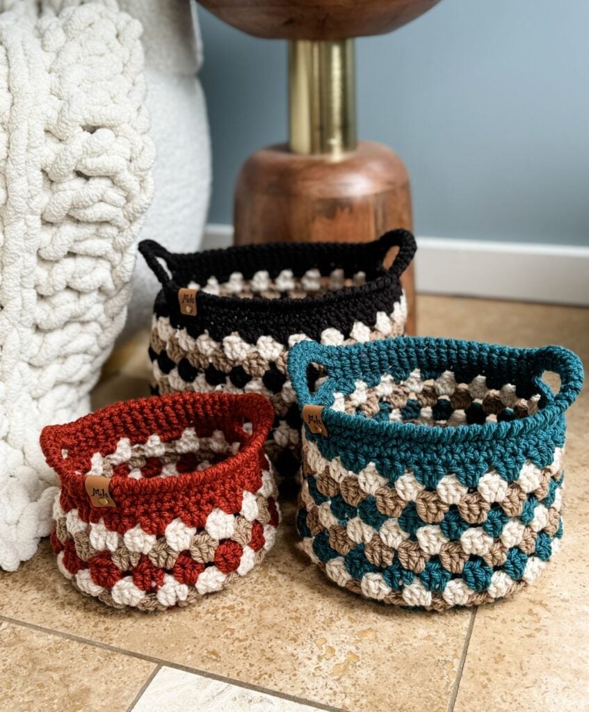 Three Granny Stitch Baskets, each in different colors (black, blue, and red) with beige accents, are placed on a tiled floor in front of a cream-colored knitted blanket.