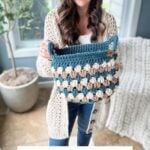 A woman in a cream sweater holds a crocheted Granny Stitch Basket with a blue and beige pattern. Text at the bottom reads, "FALL FOR GRANNY BASKETS Crochet pattern + Video.