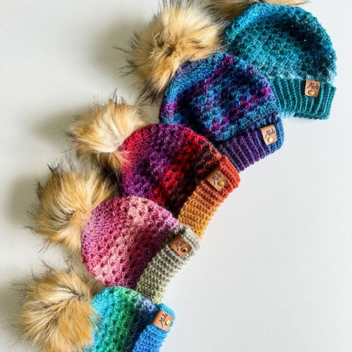 Five knitted hats in different colors, each with a faux fur pompom on top, are lined up diagonally on a white surface. Each Granny Stitch Hat features a small leather tag.
