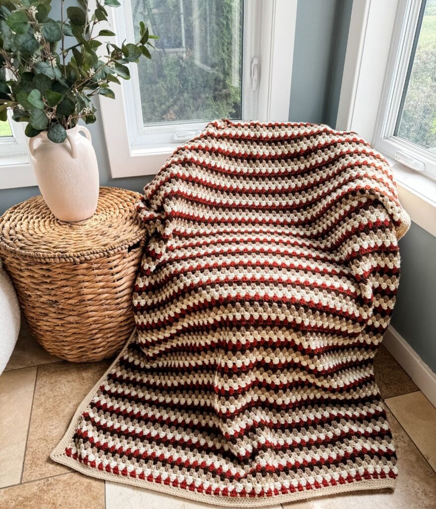 A Granny Stitch Blanket draped over a chair next to a wicker basket and a white vase with green foliage, positioned in front of large windows.