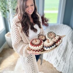 A woman holds a tray with crocheted pumpkins in a bright room with a large window and plants. She is wearing a beige crocheted sweater using the Granny Stitch over a white top, showcasing her love for crochet patterns.