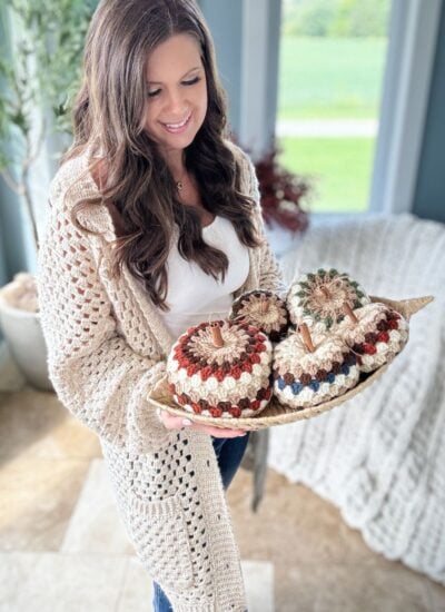 A woman holds a tray with crocheted pumpkins in a bright room with a large window and plants. She is wearing a beige crocheted sweater using the Granny Stitch over a white top, showcasing her love for crochet patterns.