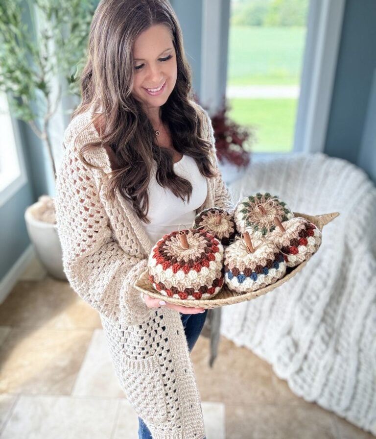 A woman holds a tray with crocheted pumpkins in a bright room with a large window and plants. She is wearing a beige crocheted sweater using the Granny Stitch over a white top, showcasing her love for crochet patterns.