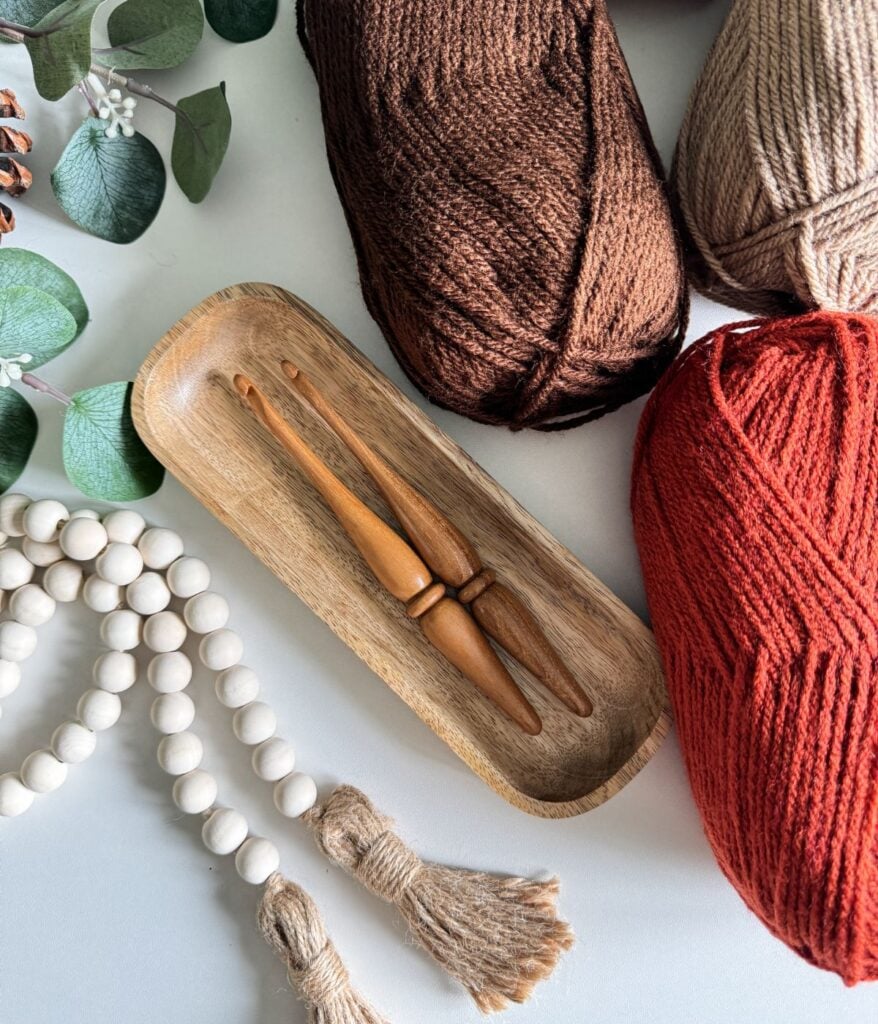 A flat lay of yarn balls in brown, tan, and red, two wooden crochet hooks in a wooden tray, green leaves, and a beaded tassel decoration on a white surface—all set for creating your next Granny Stitch Blanket.
