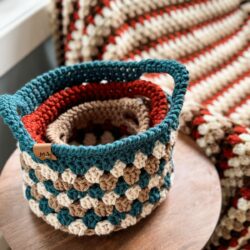 A set of intricately crocheted baskets, each differing in size and color, are nested together on a wooden surface with a matching Granny Stitch Basket blanket in the background.