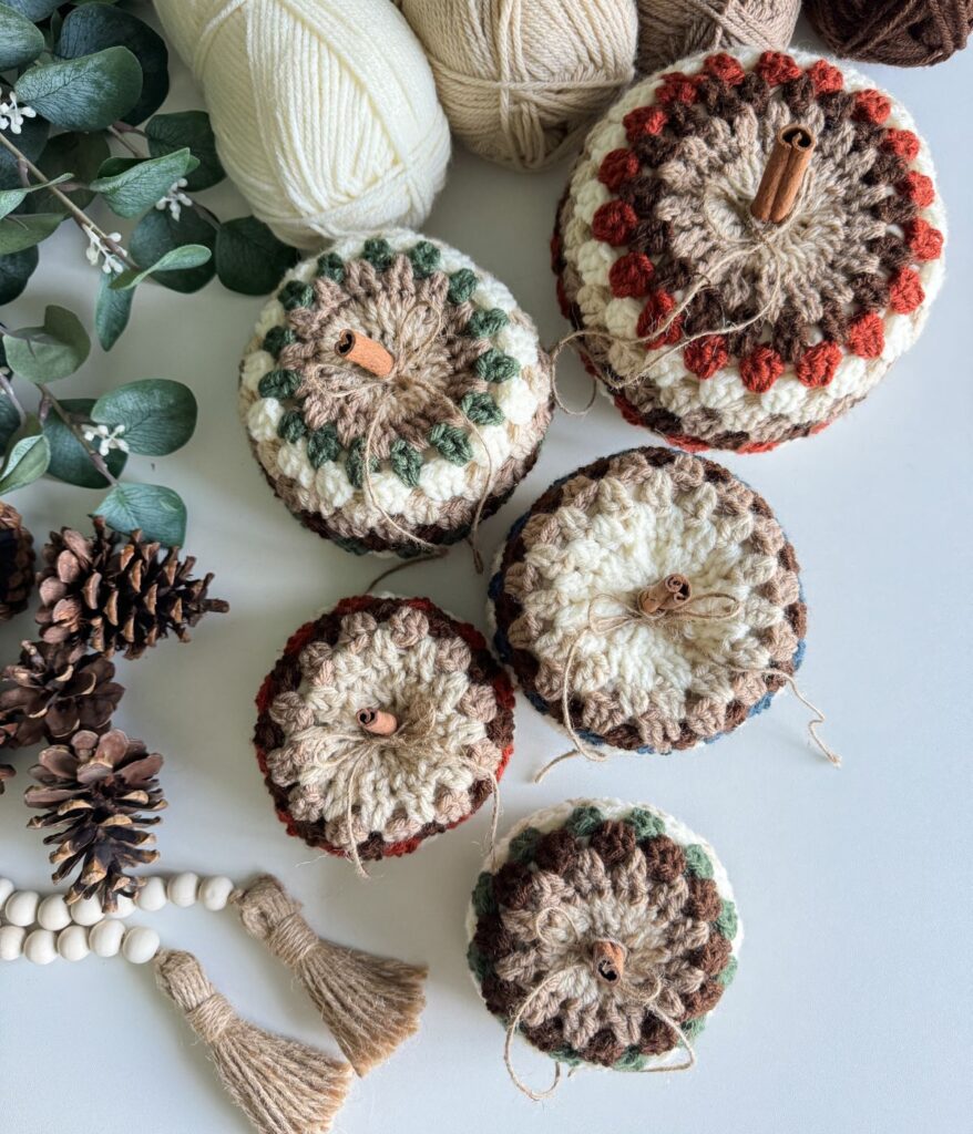 A display of five Granny Stitch Pumpkins in cozy neutral and earth tones, surrounded by yarn balls, pinecones, eucalyptus branches, wooden beads, and twine-tied tassels—a perfect touch for your fall crochet décor.