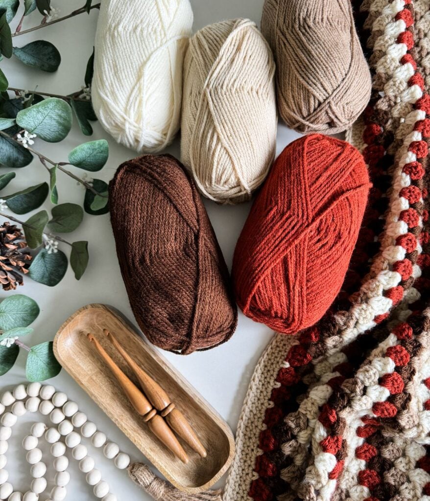 Five skeins of yarn in white, beige, brown, and red shades, two wooden crochet hooks, a crocheted Granny Stitch Blanket, a wooden tray, a string of beads, and eucalyptus leaves are arranged on a white surface.