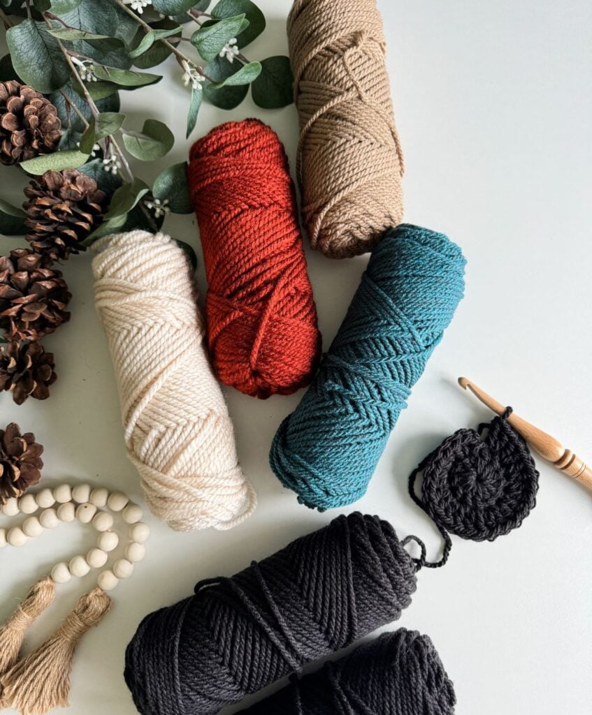 Various colors of yarn, a wooden crochet hook, pinecones, wooden beads, and greenery arranged on a flat surface alongside a charming Granny Stitch Basket.