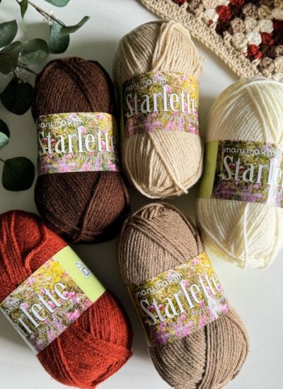 Five skeins of Mary Maxim Starlette yarn in shades of white, beige, brown, and red are displayed on a surface with greenery and a crocheted Granny Stitch Blanket in the background.