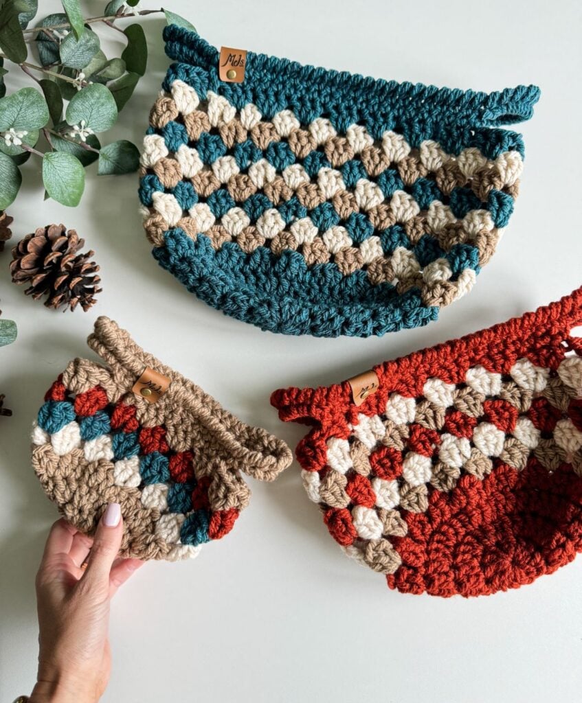 Three crocheted pouches in different sizes and colors, with a hand holding the smallest pouch. Pine cones and eucalyptus leaves are placed next to the intricately designed Granny Stitch Basket pouches.