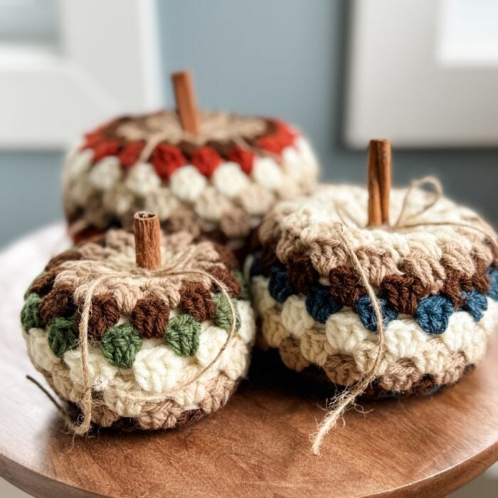 Three handmade crochet pumpkins, featuring cinnamon stick stems and twine bow details, rest on a wooden surface near a window. Perfect for fall decor, these charming pumpkins can be crafted using a free crochet pattern with the classic granny stitch pumpkin design.