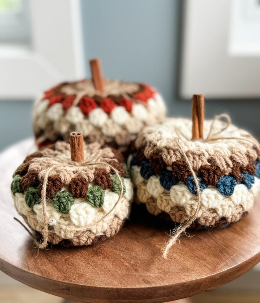 Three handmade crochet pumpkins, featuring cinnamon stick stems and twine bow details, rest on a wooden surface near a window. Perfect for fall decor, these charming pumpkins can be crafted using a free crochet pattern with the classic granny stitch pumpkin design.