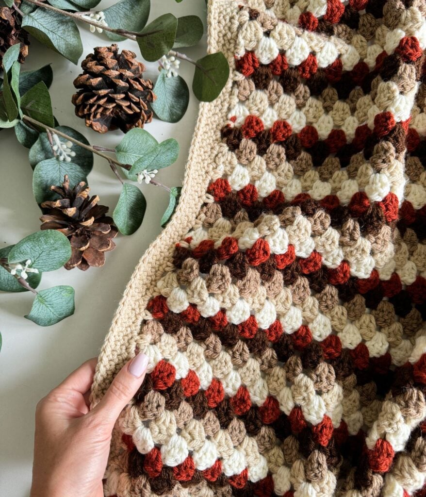 A person holds a multicolored Granny Stitch Blanket. Pinecones and eucalyptus leaves lie beside the blanket on a white surface.