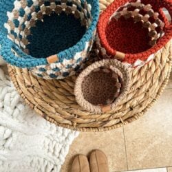 Three crochet baskets in blue, red, and brown, crafted with a charming granny stitch, are placed on a woven round side table next to a white textured blanket. Brown slippers are visible at the bottom of the image.