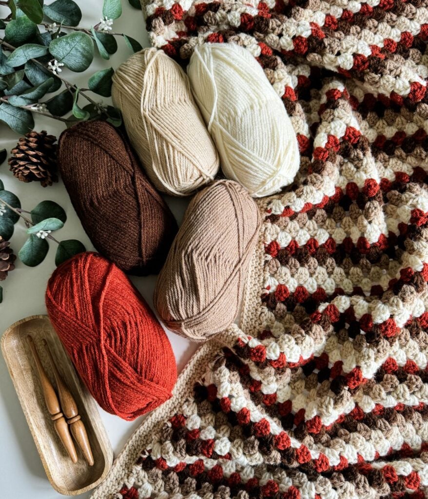 Five skeins of yarn in cream, white, brown, beige, and red sit beside a matching Granny Stitch Blanket, wooden crochet hooks, pine cones, and eucalyptus leaves.