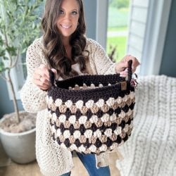 A woman in a cream sweater holds up a black and tan Granny Stitch Basket in a bright room with plants and a window in the background.