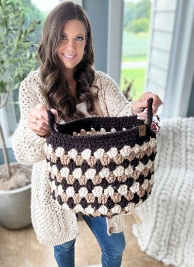 A woman in a cream sweater holds up a black and tan Granny Stitch Basket in a bright room with plants and a window in the background.