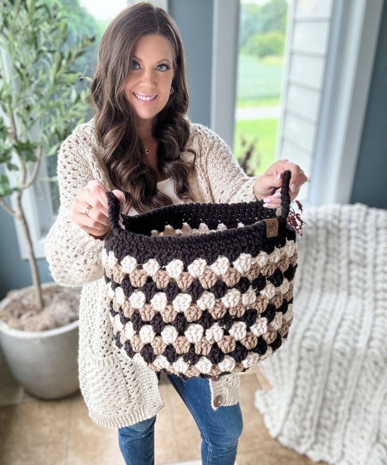 A woman in a cream sweater holds up a black and tan Granny Stitch Basket in a bright room with plants and a window in the background.