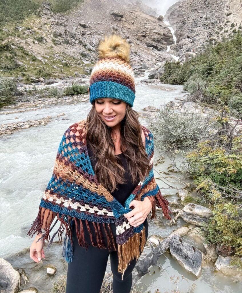 A woman wearing a Granny Stitch Hat and shawl stands by a rocky river with a mountainous landscape in the background.