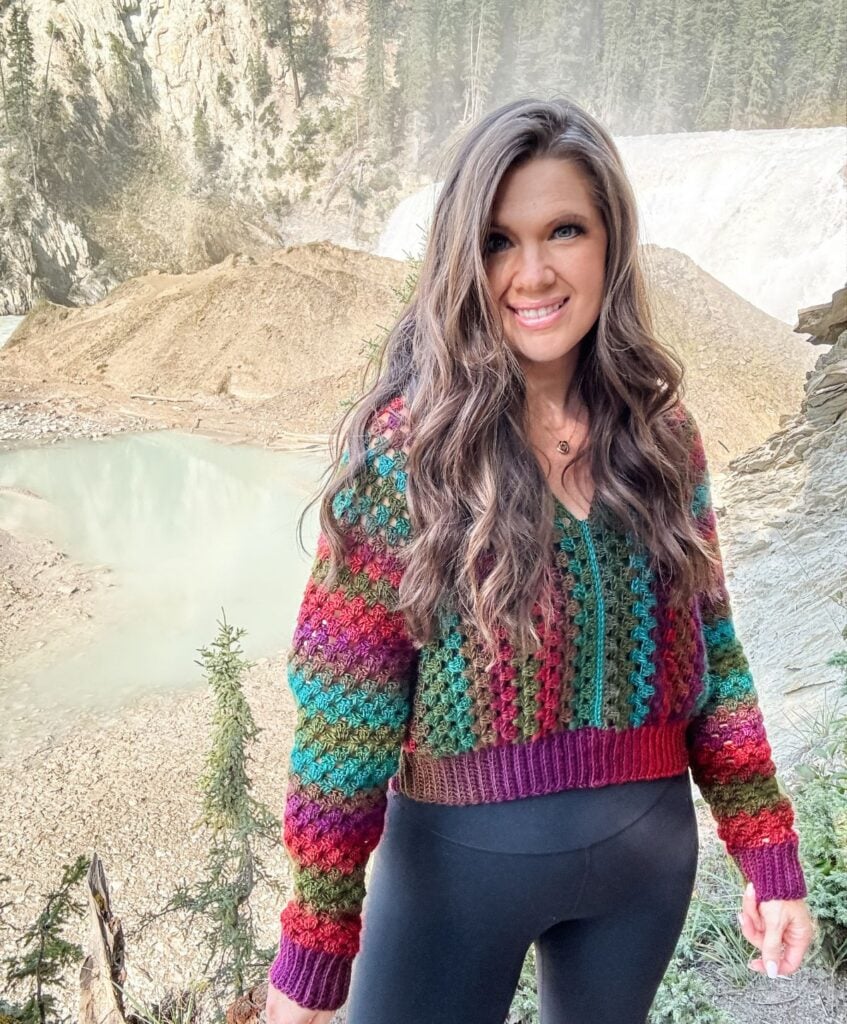 A woman with long hair stands outdoors in front of a mountainous landscape with a body of water. She is wearing a colorful crochet pullover and black leggings, smiling at the camera.