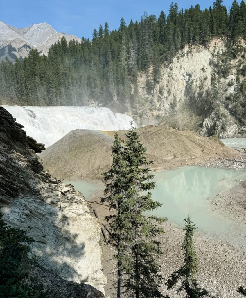 A waterfall cascades down a rocky cliff into a pool of milky turquoise water, surrounded by trees and mountains in the background, creating a scene as cozy and inviting as your favorite crochet pullover.