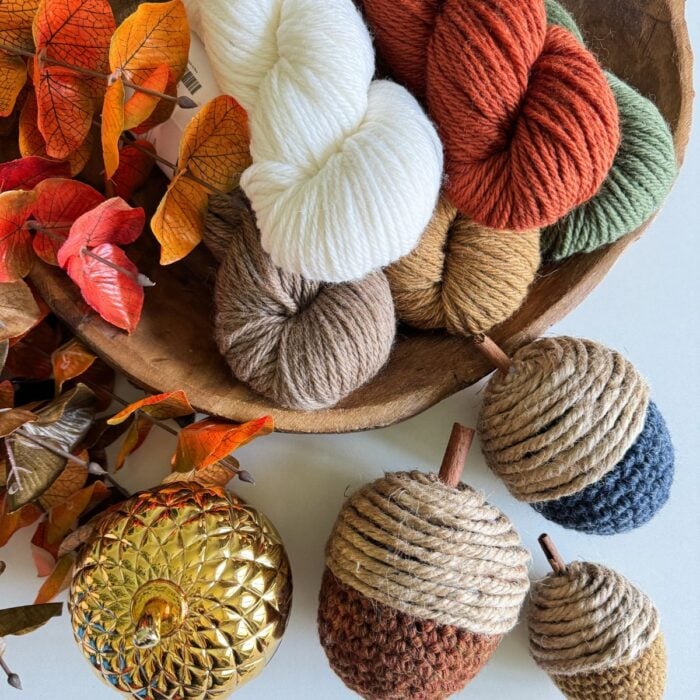 A wooden bowl holds skeins of yarn in autumn colors, among which are crochet acorns and a metallic pumpkin. Orange and red leaves are placed to the side.