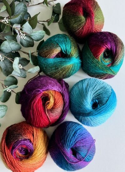 Balls of multicolored yarn are arranged beside fresh green eucalyptus leaves on a light surface, inspiring visions of a cozy crochet cardigan.
