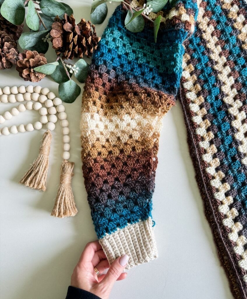Hand holding knitted sleeve with multicolored pattern; decorative beads, tassels, pinecones, and green leaves are also on the table. A cozy crochet cardigan rests nearby, adding to the charming scene.