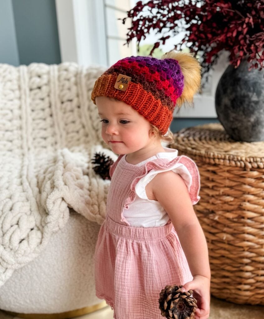 A toddler wearing a colorful Granny Stitch Hat and a pink dress holds pinecones, standing in a cozy, decorated room with a woven basket and a chunky blanket in the background.