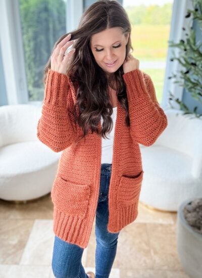 A woman with long, dark hair in a cozy, orange knit cardigan, touches her hair while standing in a bright room with white chairs and a potted plant.