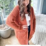 Woman wearing a fuzzy peach cardigan stands indoors next to a potted plant and white knit blanket. Text reads: "Fuzzy Peach Cardigan. Crochet pattern + Video.