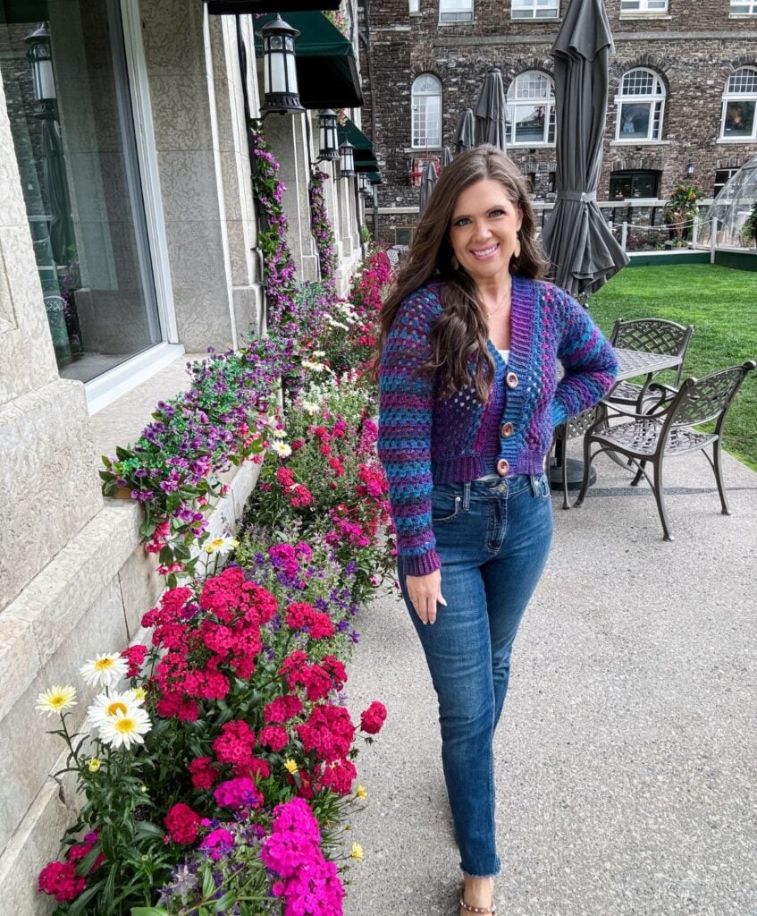 A woman stands on a walkway beside a building adorned with a variety of colorful flowers. She is wearing jeans and a multi-colored crochet cardigan, smiling at the camera.