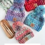 Four colorful Granny Stitch Hat crochet hats with pom-poms are displayed next to two crochet hooks and a wooden tray on a white surface. Text below reads "Granny Stitch Hat Crochet Pattern & Video.
