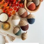 Decorative crochet acorns in various colors displayed with autumn leaves, a candle, beads, and a wooden bowl, alongside a sign reading "Rustic Farmhouse Acorns Crochet pattern + Video.