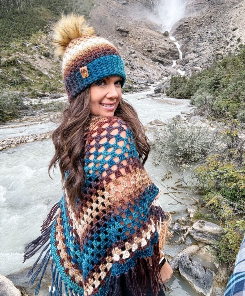 A woman in colorful knit attire and a charming Granny Stitch Hat smiles while standing near a river with a mountainous backdrop.