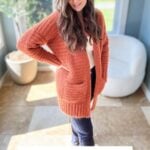 A woman wearing an orange knitted cardigan stands indoors by a window, with hands on hips. Text overlay reads, "Fuzzy Peach Cardigan, Crochet pattern + Video.