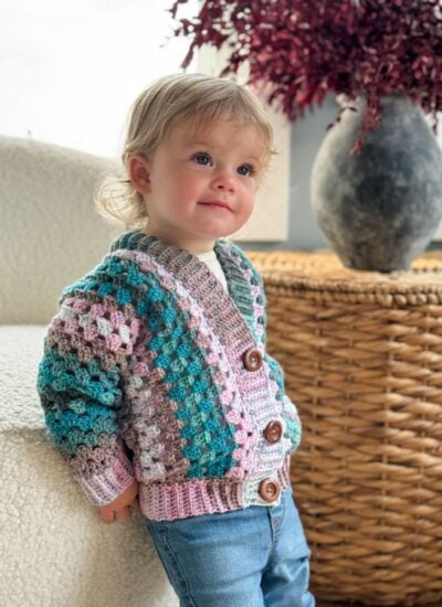 A young child wearing a vibrant Children's Crochet Cardigan and jeans leans against a white chair. In the background, there is a wicker basket with a gray vase and red foliage.