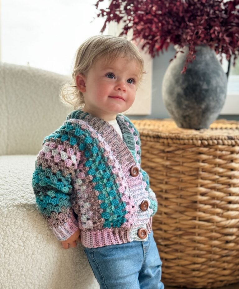 A young child wearing a vibrant Children's Crochet Cardigan and jeans leans against a white chair. In the background, there is a wicker basket with a gray vase and red foliage.