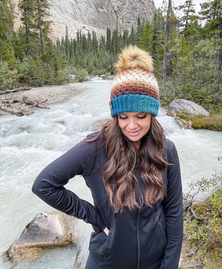 A woman wearing a multicolored Granny Stitch Hat and a black jacket stands by a flowing river with a forest and mountain in the background.