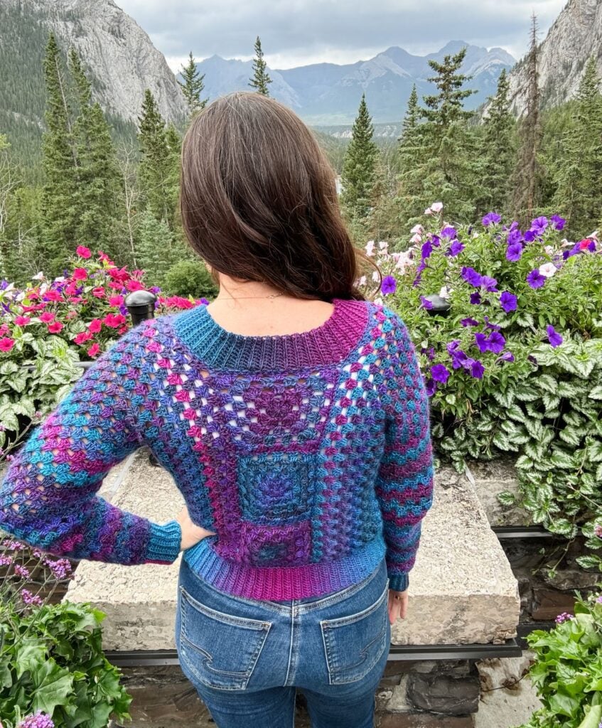 A person with long dark hair stands with their back to the camera, wearing a colorful crochet cardigan and jeans. They are in a mountainous area surrounded by vibrant flowers and evergreen trees.