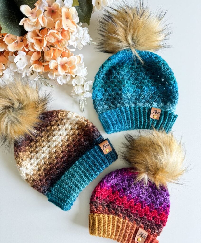 Three Granny Stitch Hats with pom-poms are laid out on a white surface next to a bunch of flowers. The beanies are multicolored, in shades of blue, brown, and red.
