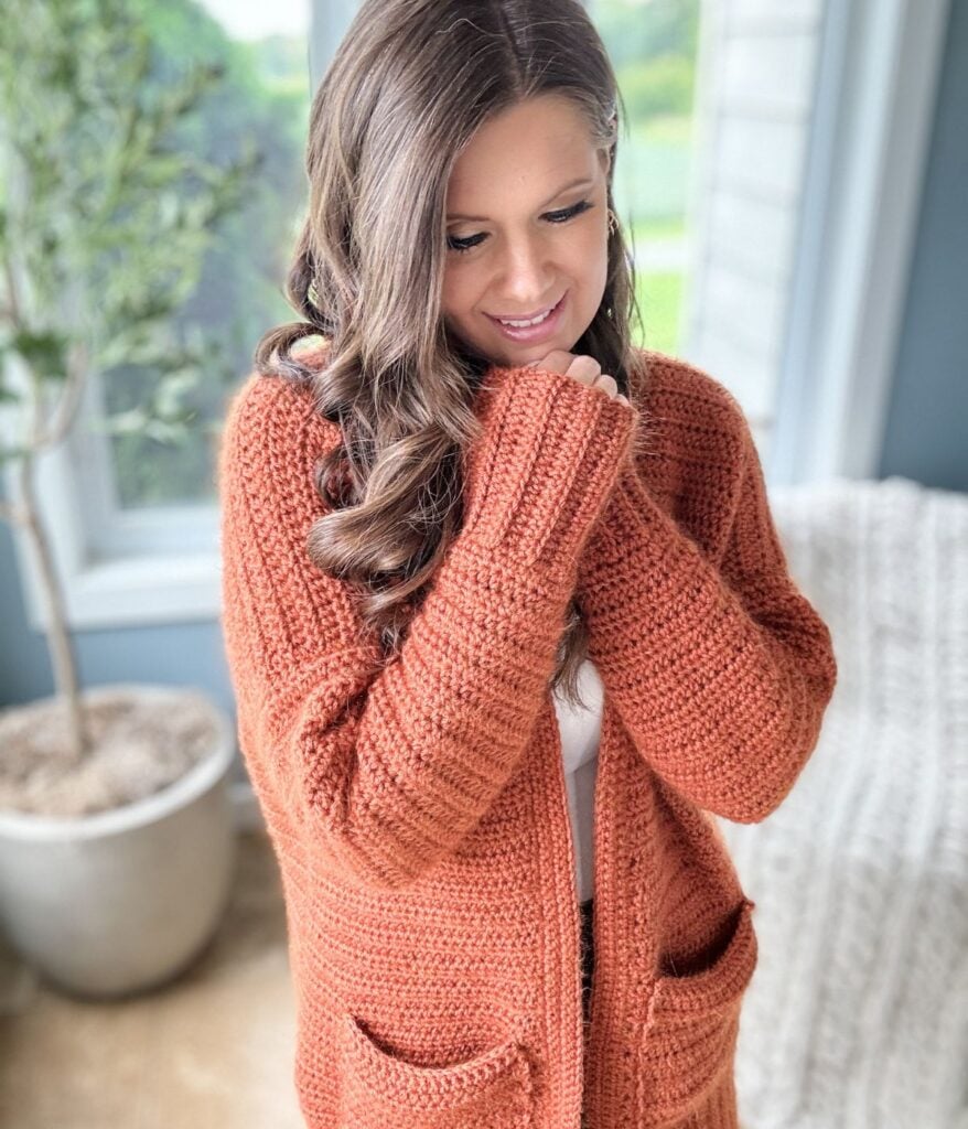A woman stands indoors wearing an orange knit sweater, looking down and smiling, with one hand touching her sweater. A potted plant and window are in the background.