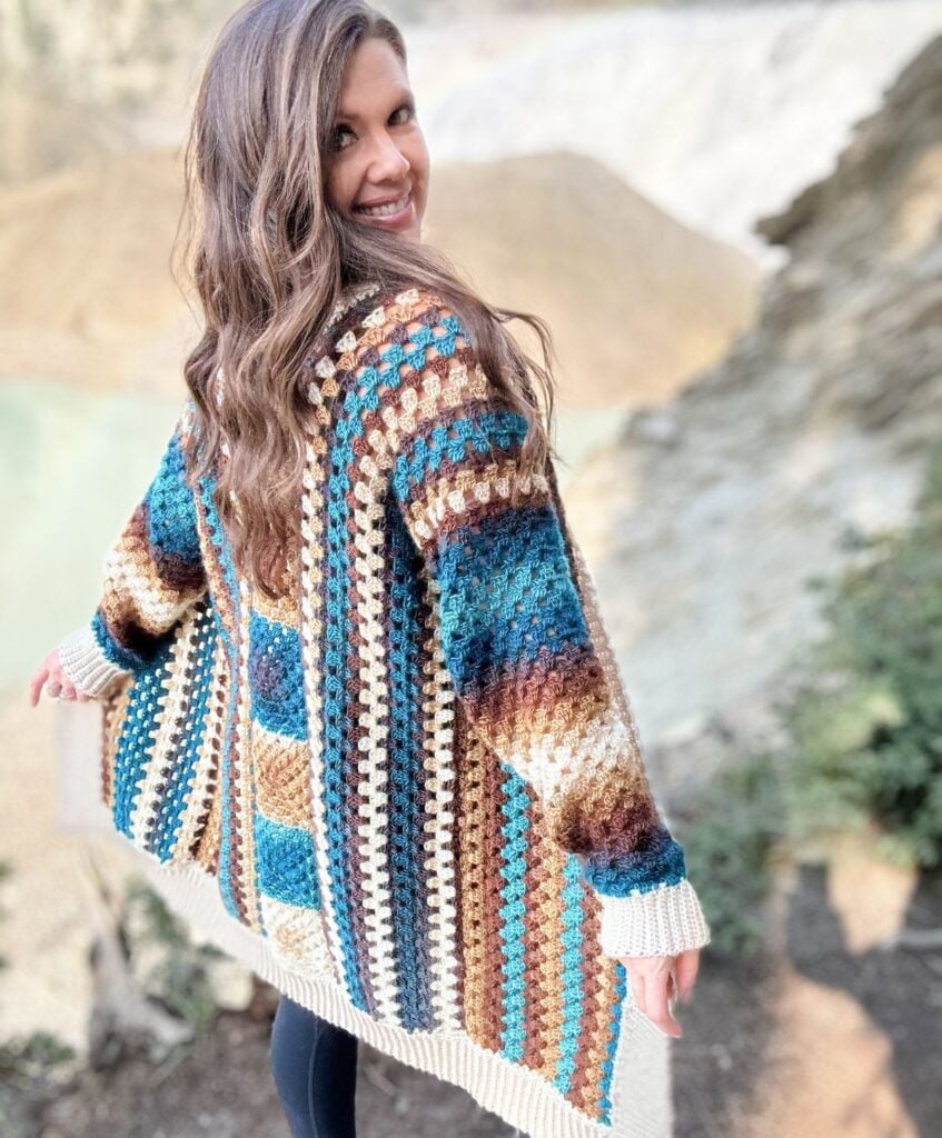 A person with long brown hair is standing outdoors, wearing a colorful, crochet cardigan. They are turned slightly, looking back over their shoulder and smiling.