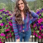 A woman stands in front of a mountain landscape and vibrant flowers, wearing a colorful crochet cardigan. Text at the bottom reads, "Cascade Cardigan Crochet Pattern, XS-5X + Video.