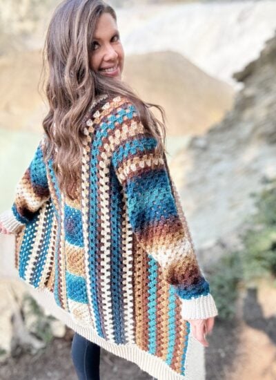 A person with long hair smiles and looks over their shoulder, wearing a crochet cardigan, standing outdoors with a rocky and green landscape in the background.
