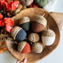 A wooden bowl holds a variety of large crochet acorns in different colors, surrounded by yarn and autumn leaves. A hand is visible at the bottom of the image.