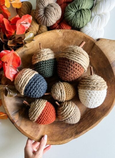 A wooden bowl holds a variety of large crochet acorns in different colors, surrounded by yarn and autumn leaves. A hand is visible at the bottom of the image.