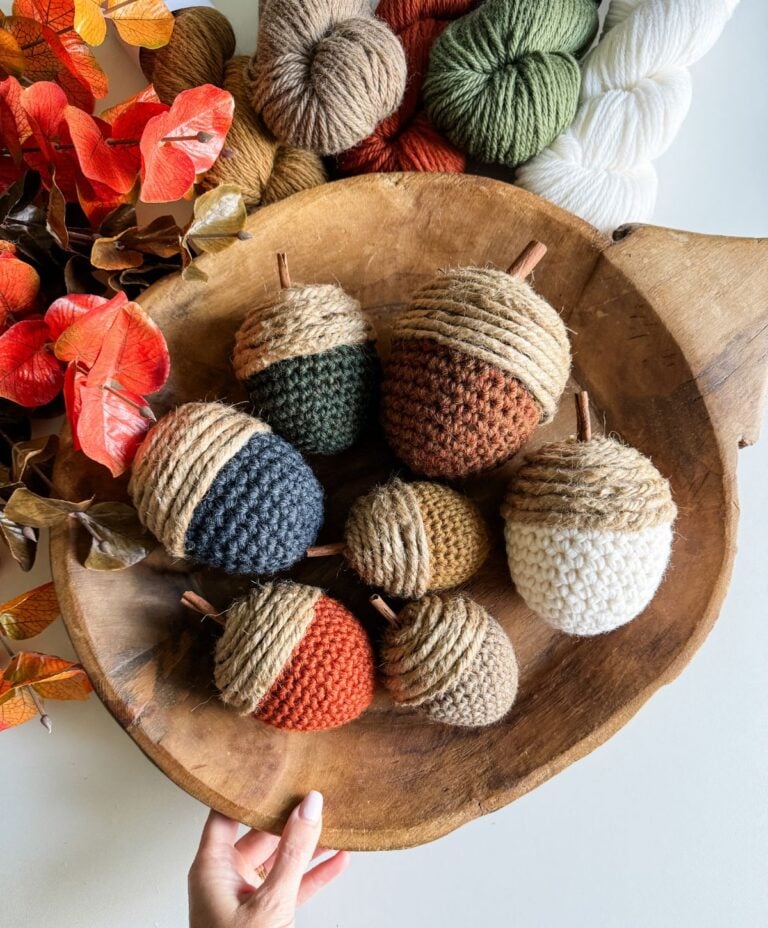 A wooden bowl holds a variety of large crochet acorns in different colors, surrounded by yarn and autumn leaves. A hand is visible at the bottom of the image.