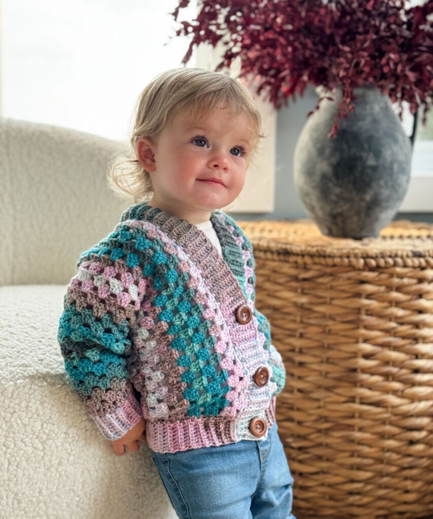 A toddler wearing a vibrant crochet cardigan and jeans stands indoors with a woven basket and a vase of red foliage in the background.